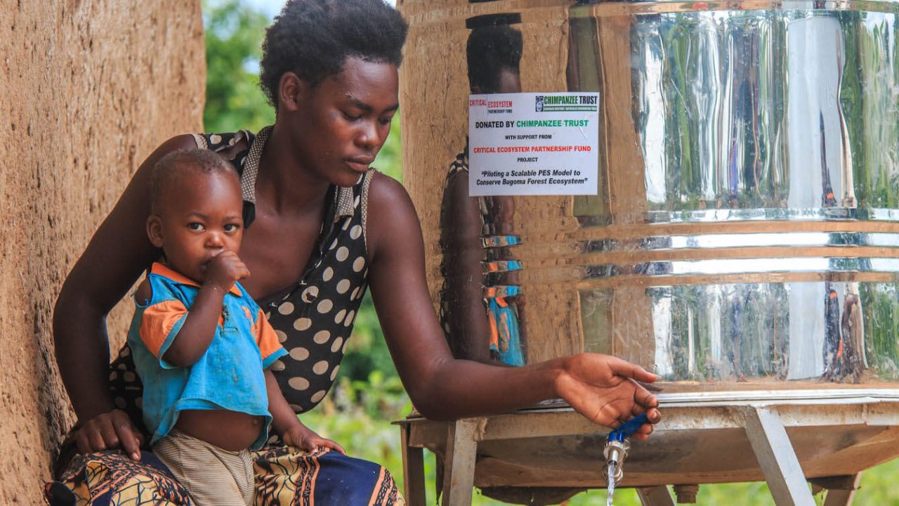 Woman with young child opens a metal drum's faucet, dispensing water.