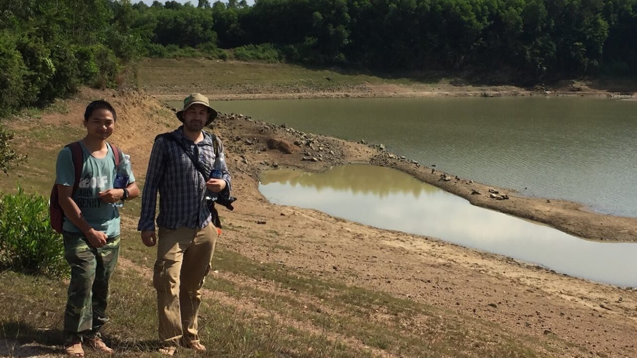 Two men standing by dark-green body of water.