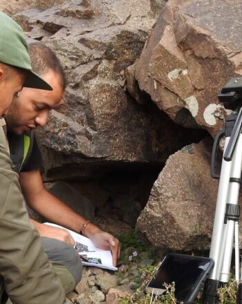 Two men crouching down near boulders, examining papers.