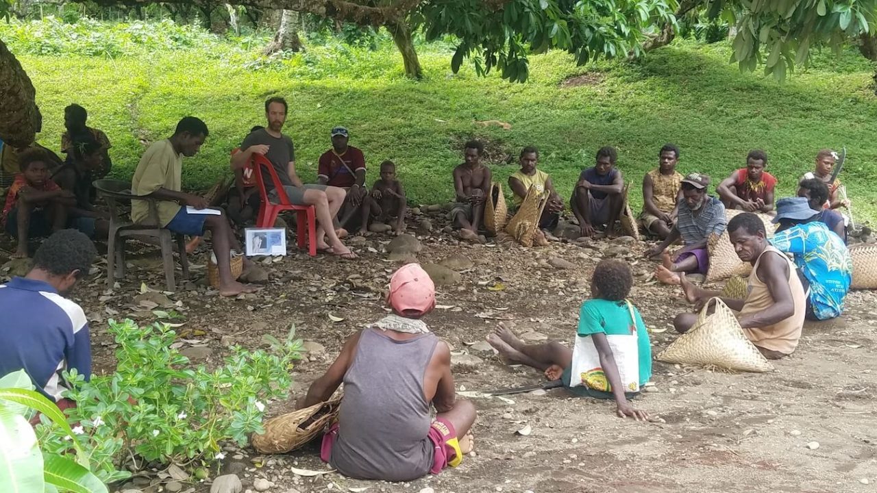 About 20 people sitting outside in a circle, under a tree.