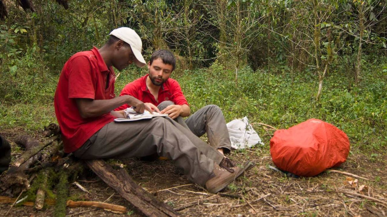 Two men sitting on forest floor, looking at notebook.