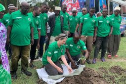 A crowd of people gathers behind a woman and man planting a tree seedling in the ground.