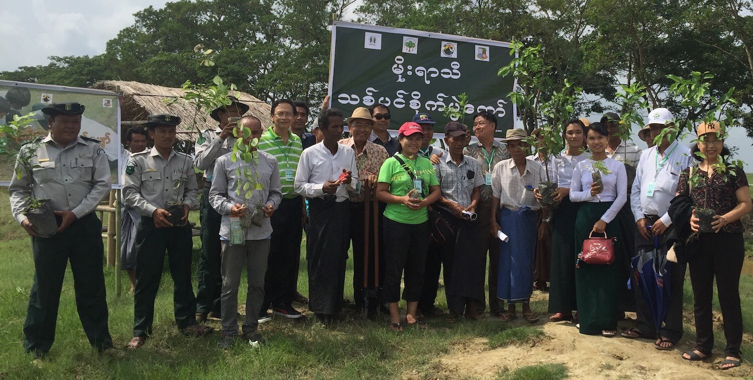 Group of about 20 people smiling, each holding a tree sapling.