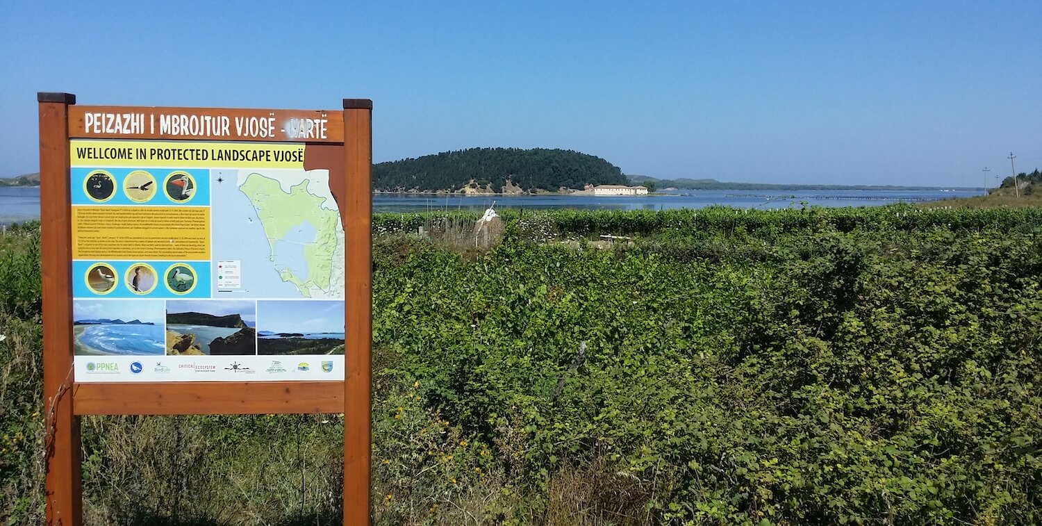 Protectee landscape sign in front of greenery and, beyond that, water.