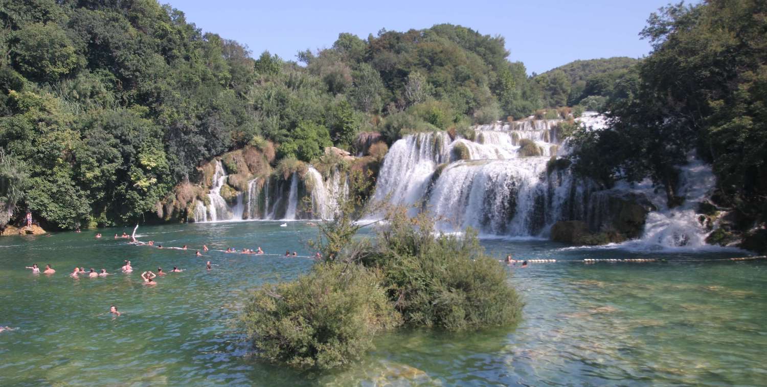 People swimming in emerald green water, series of waterfalls in the background.