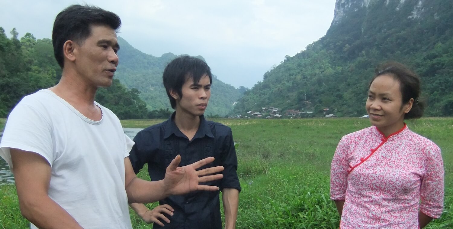 Two men and one woman having a discussion in the foreground, lush landscape in the background.