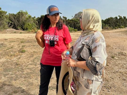 Two women converse in an open field near a wooded area.