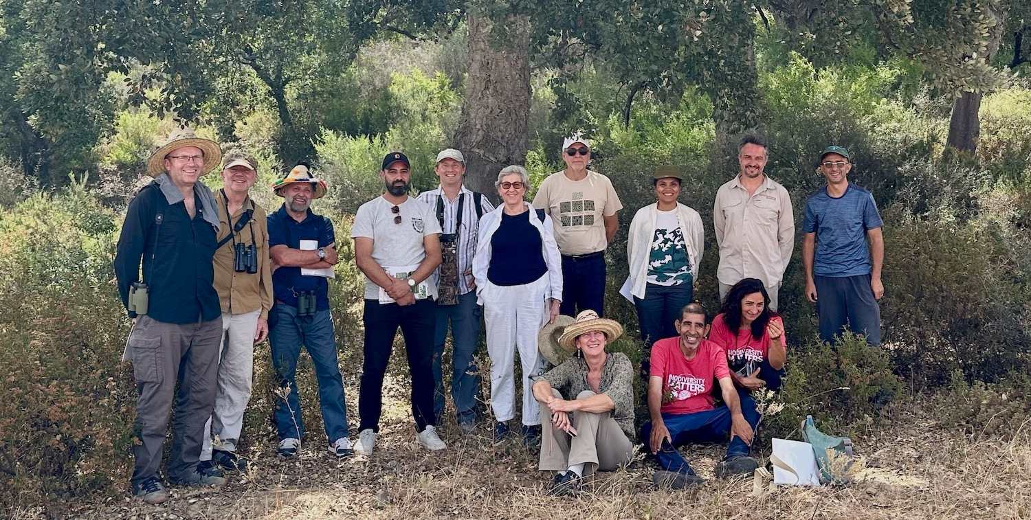 A group of women and men pose for a group photo in a wooded area.