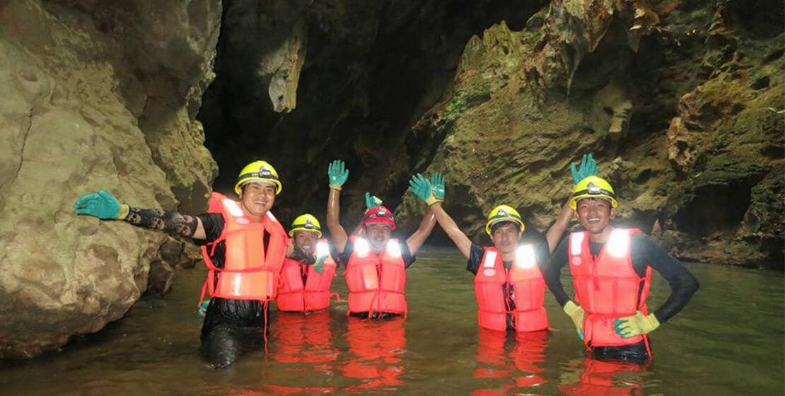 Five people wearing helmets and orange vests in cave, waist-height in water, smiling, waving at camera.