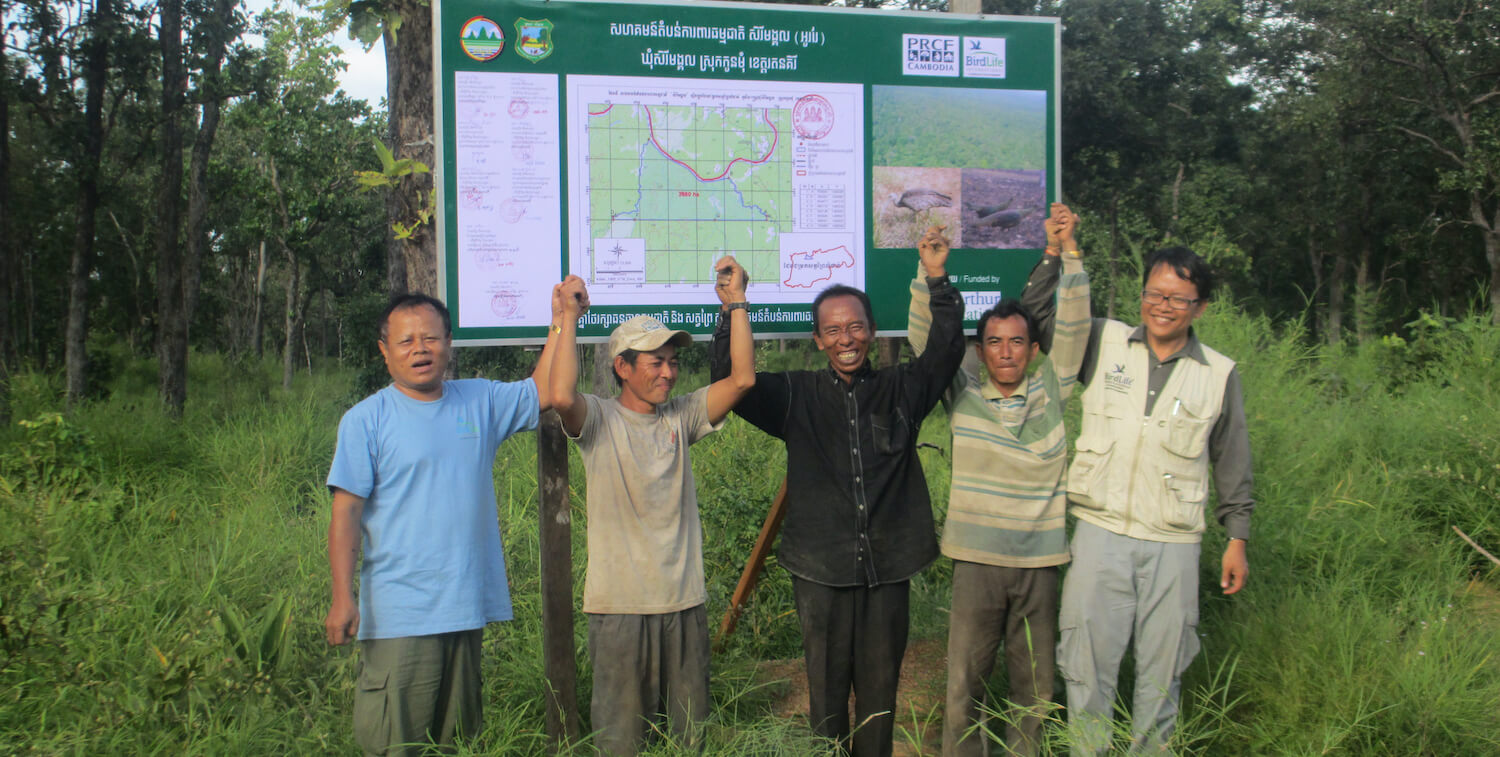 Group of 5 men standing outside, holding hands in the air and smiling in front of signboard with map.