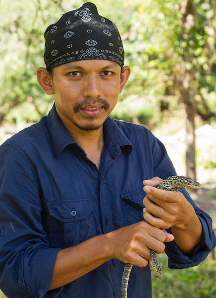 Close-up of Deni Purwandana wearing a black bandana on his head and holding a small Komodo dragon hatchling with both hands.