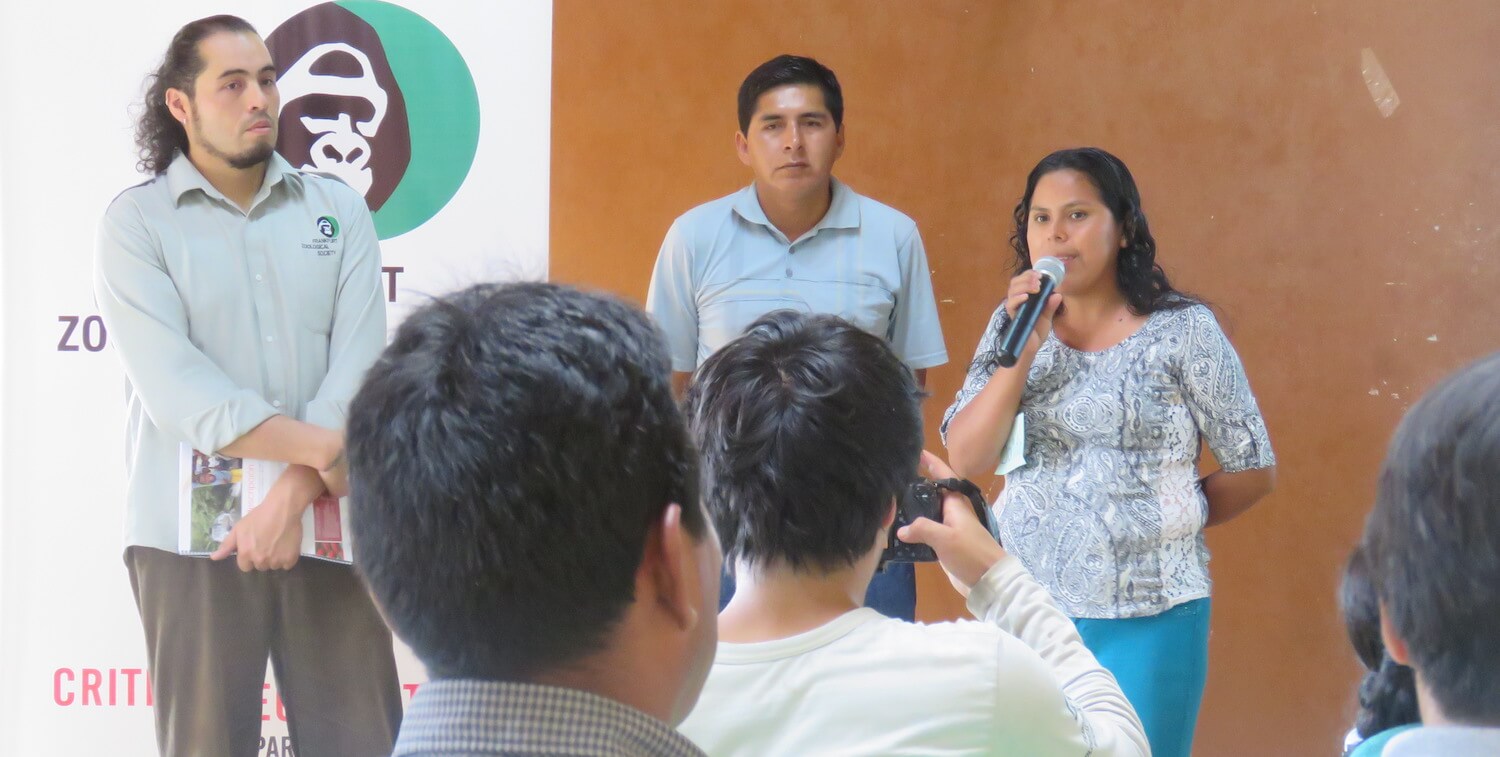 Tres personas de pie frente a un grupo, una mujer hablando por el micrófono.