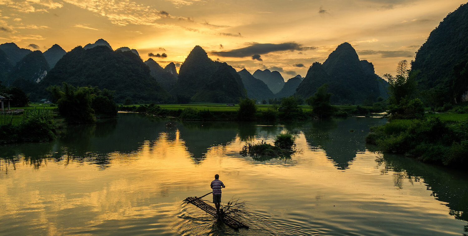 丸太でできた細い筏の上に立って、夕日が沈むにつれて水面を漕いでいる男性。