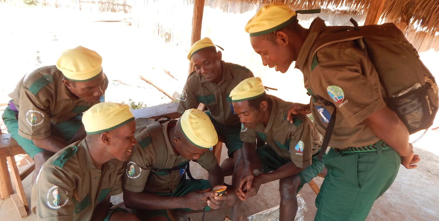 Six men in uniform, yellow caps, brown shirts and green pants, looking at small monitoring device.