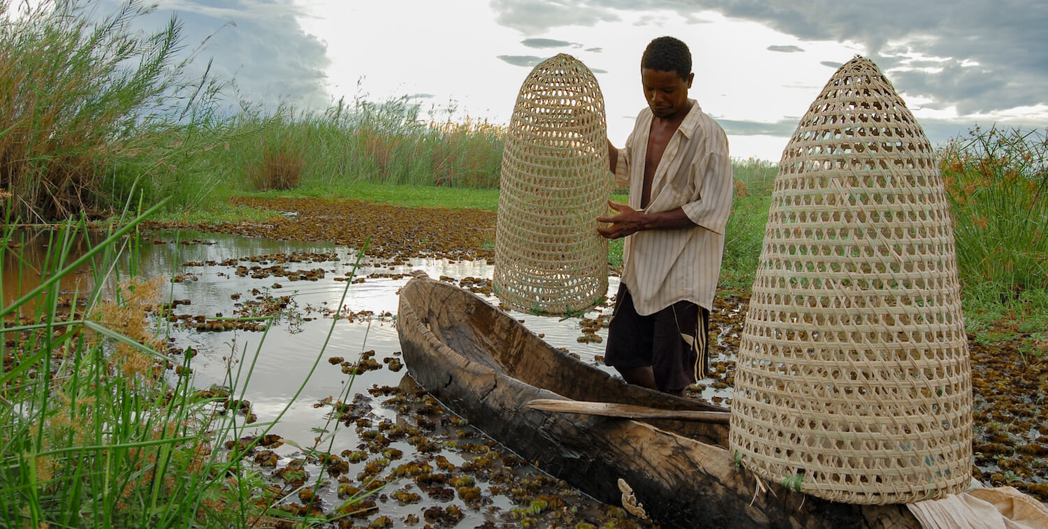Man standing next to canoe in shallow water that holds two cone-shaped fish traps.