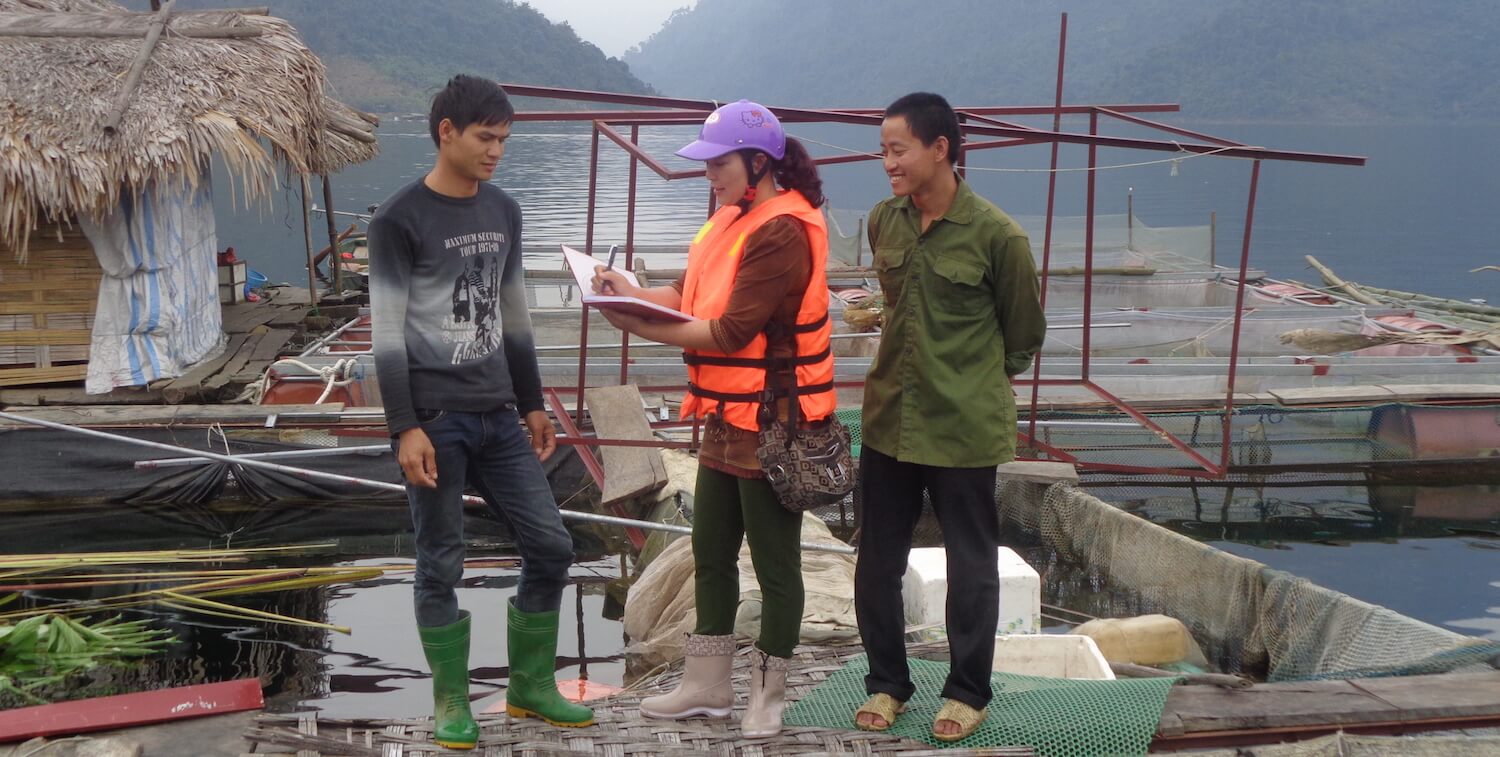 Junto al agua, dos hombres se paran junto a una mujer que está escribiendo en un cuaderno.