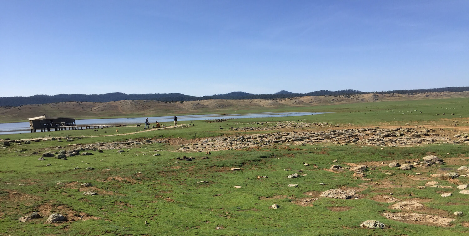 Paisaje verde y llano, pequeño edificio al lado del lago en el fondo.