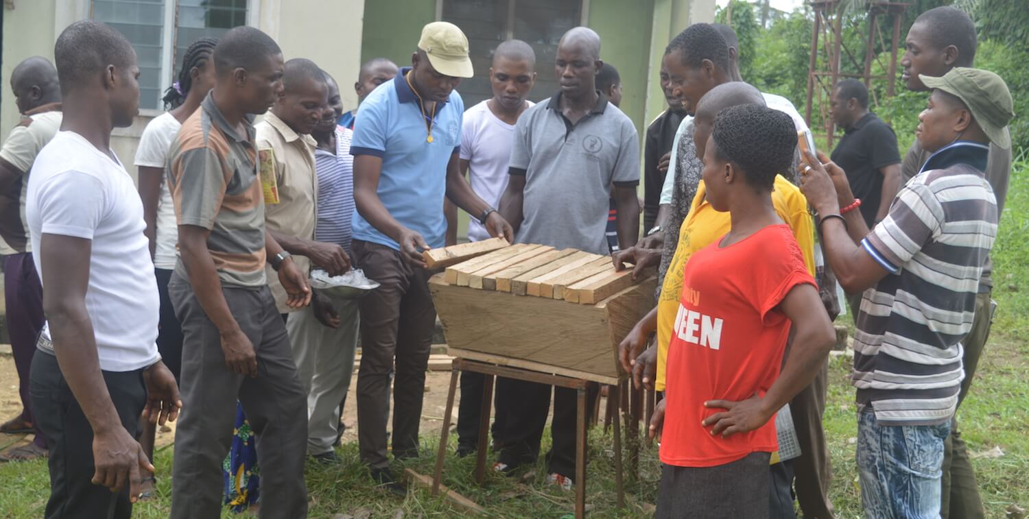 Man finishing wooden beehive while group of people around him observes.