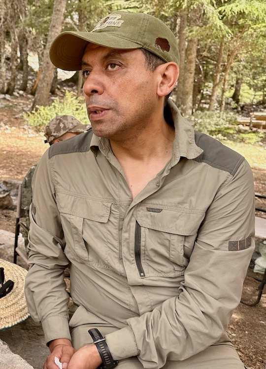 A man in a khaki shirt and ball cap sits near a wooded area.