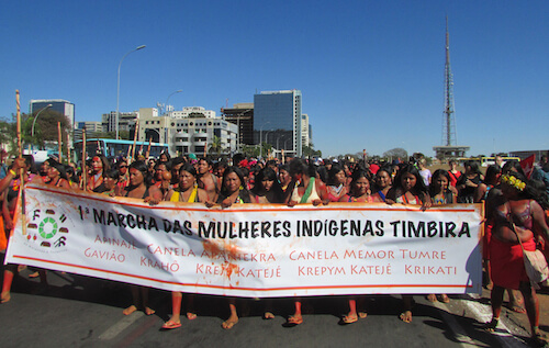 Indigenous women holding long banner, marching in the street.