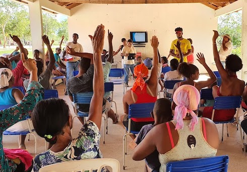 People raise hands during meeting in open-air building.