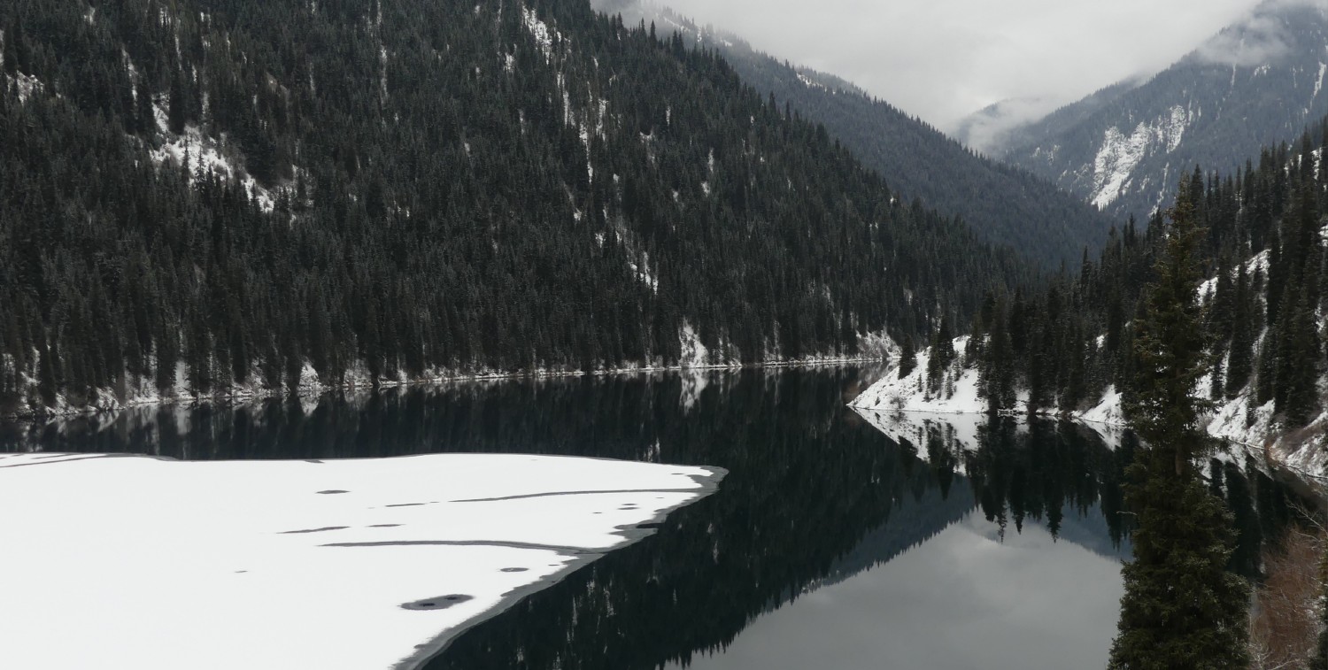 View of mountains reflecting in a dark lake with patches of snow.