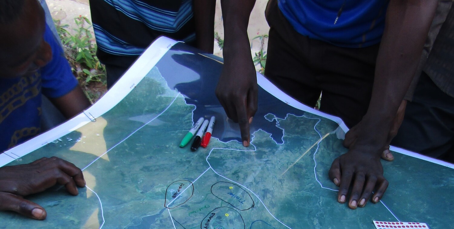 Map on table, a few people's hands holding down map, one pointing toward location..