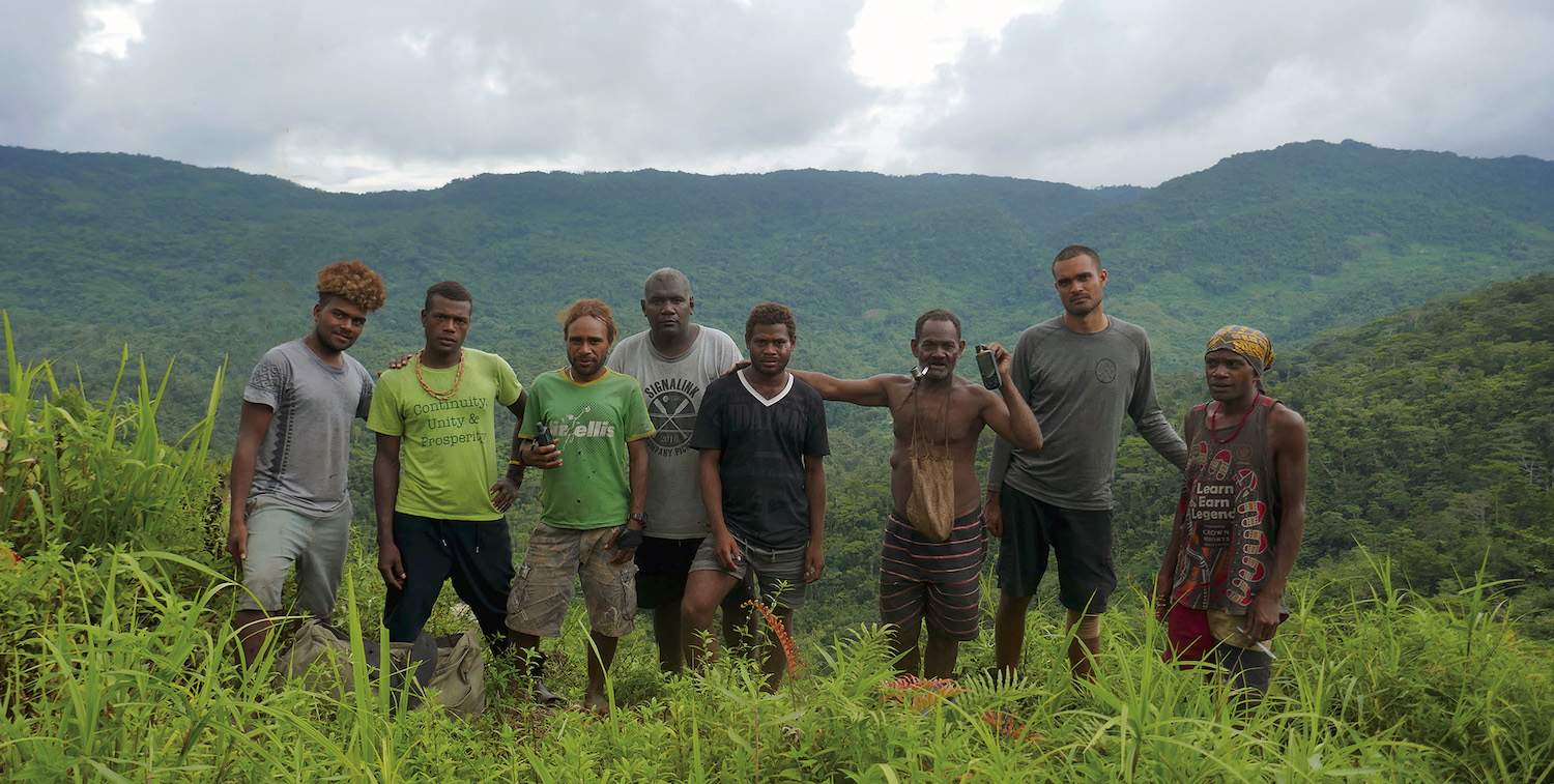 A group of men pose standing in lush greenery with mountains in the background.