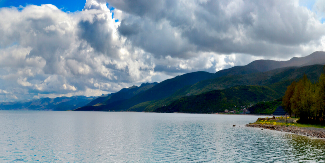 Vista del agua con exuberantes montañas de fondo, nubes y cielo azul brillante.
