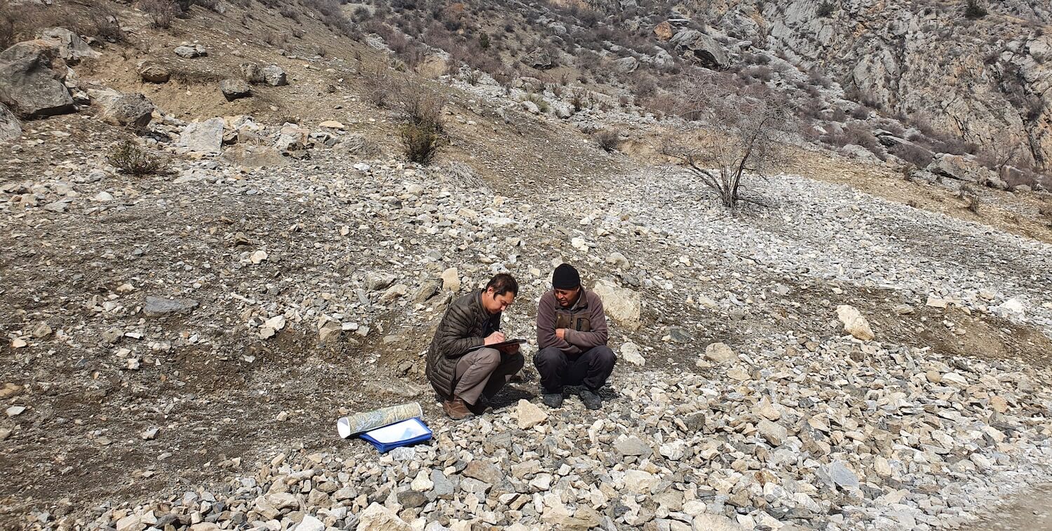 Two men in rocky landscape crouched on ground, talking.