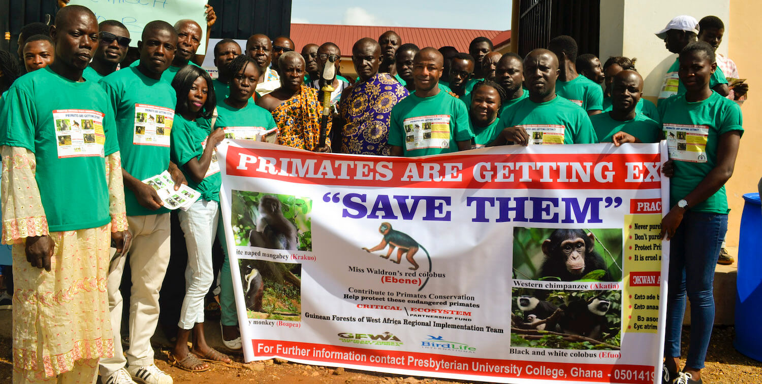 バナーの後ろに立っている緑色のシャツに、類人猿の写真と「SaveThem」と他のテキストを合わせたカップルのグループ。