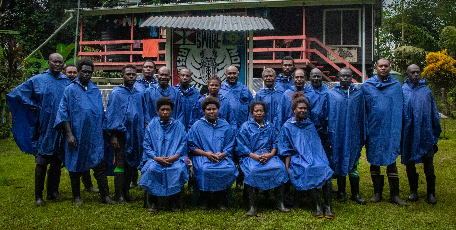 Grupo de unas 20 personas con ponchos azules para la lluvia, de pie/sentados frente a la estación de guardabosques.