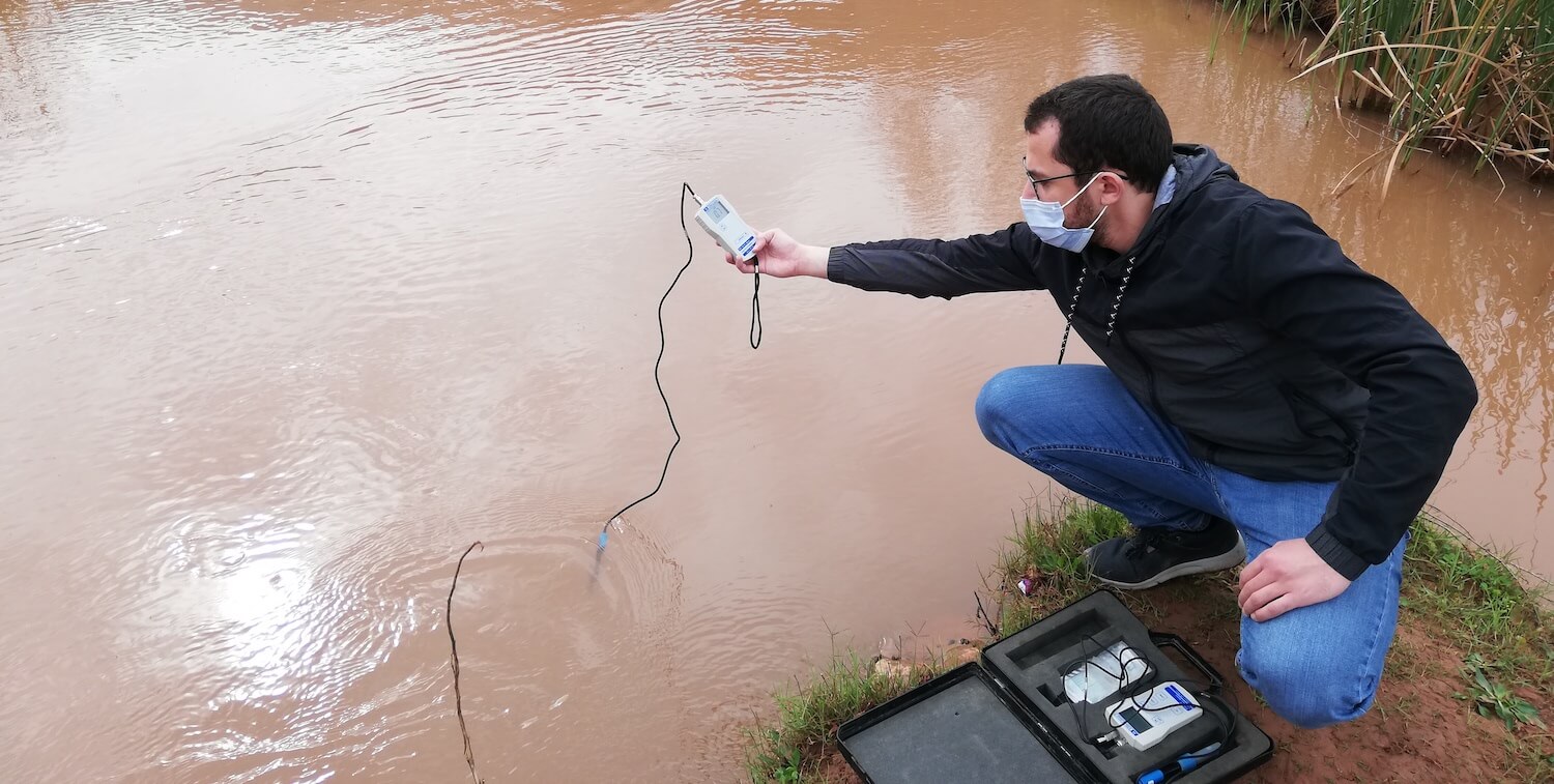 Man kneeling down near brown water with a monitoring device in his hand that goes into the water.