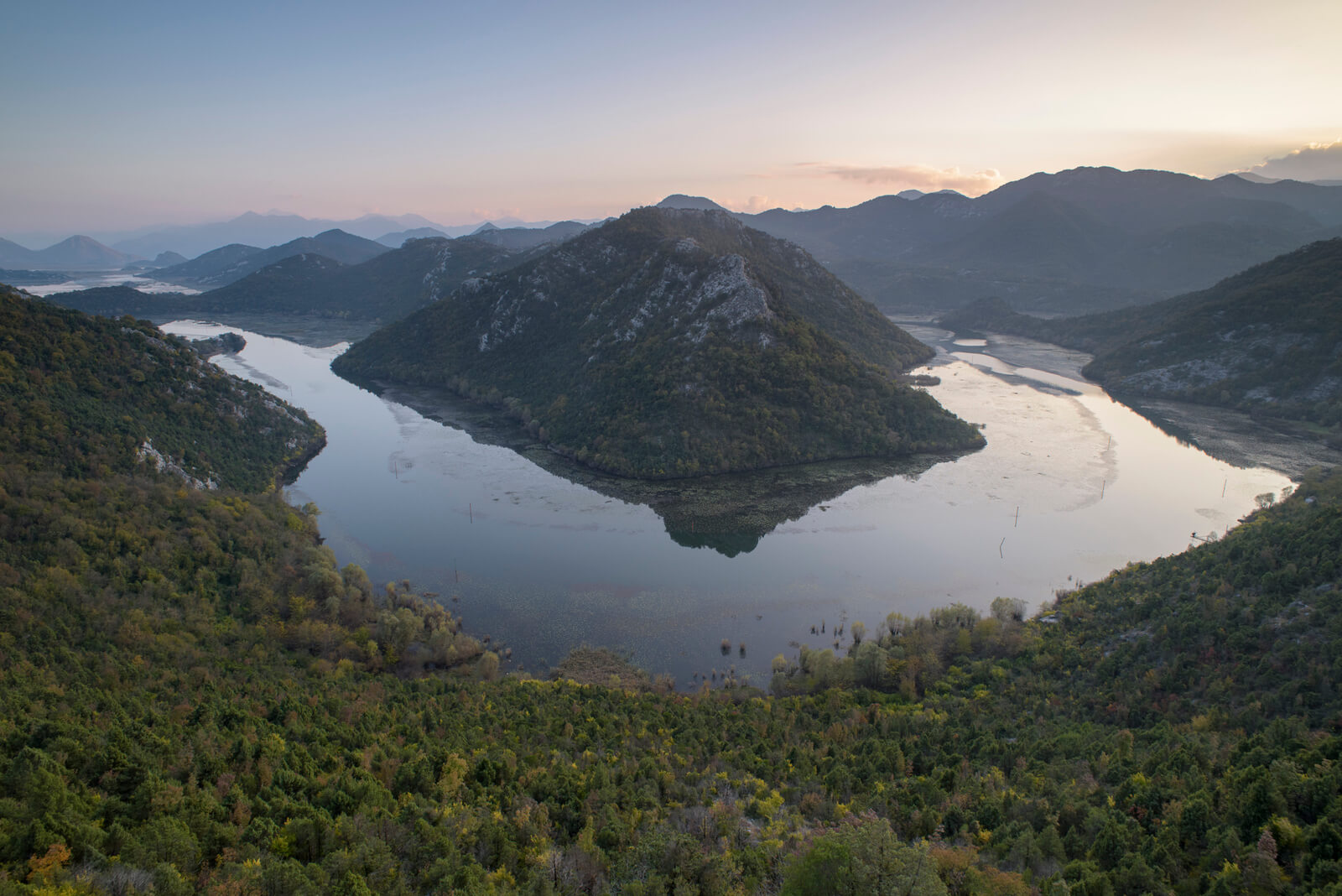 Large river circling small mountain, with more mountains in the background. Trees in foreground.