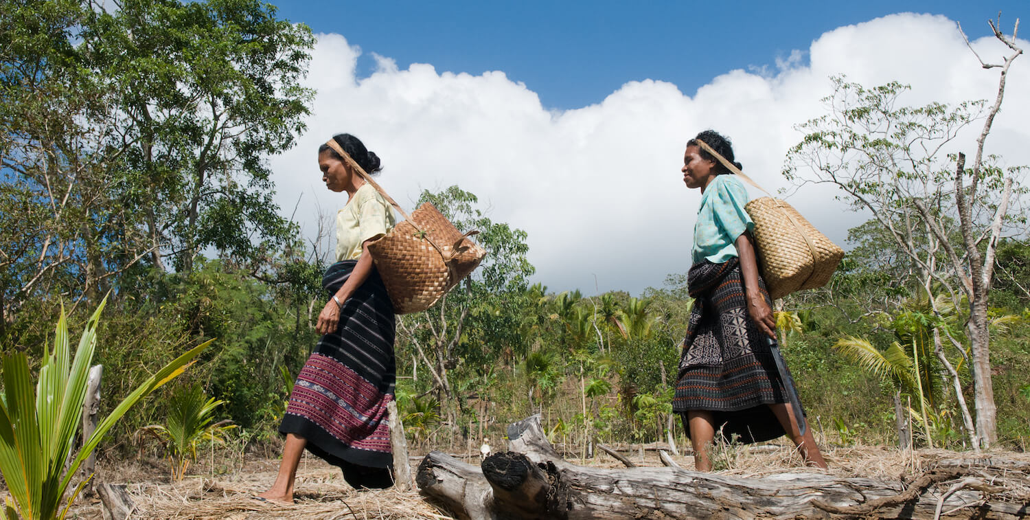 Two women in traditional dress walking outside, holding baskets with straps on head.