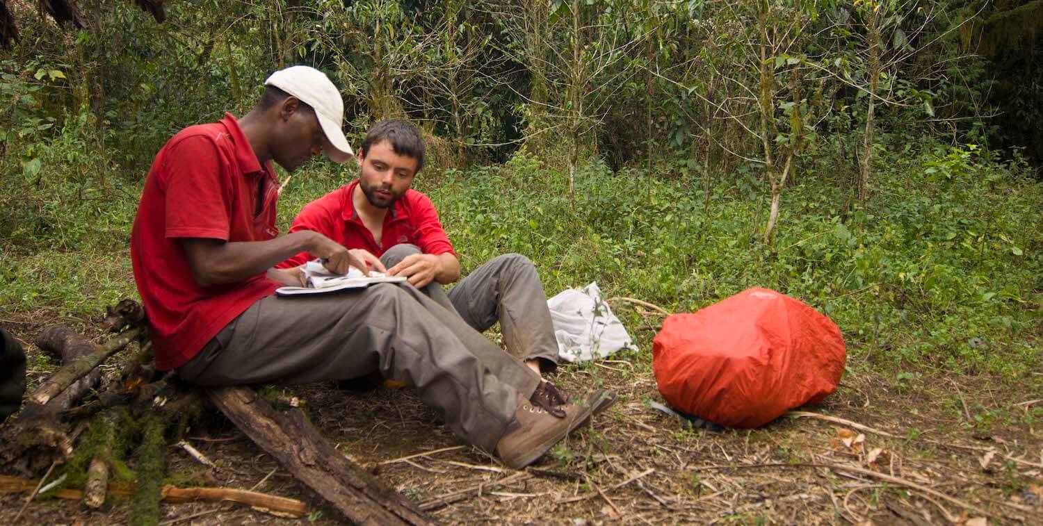 Dos hombres sentados en el suelo del bosque, mirando el cuaderno.