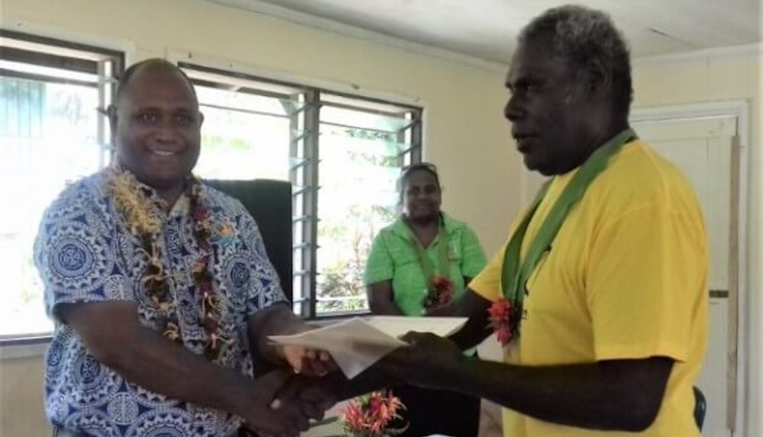 Two standing men, shaking hands while also passing a set of papers between them.
