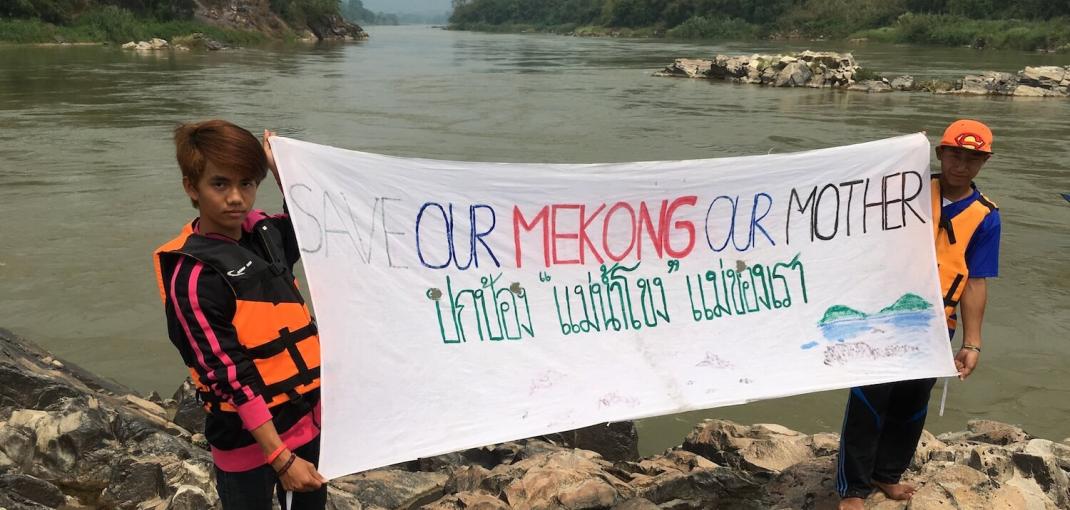 Dos niños de pie sobre rocas frente a un río muestran un cartel que apoya la conservación del río Mekong, Tailandia.