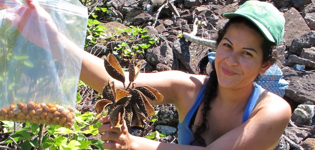 Mujer sonriente sostiene una bolsa de semillas en una mano y una planta en la otra.