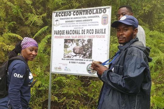 A woman and two men stand next to a sign near thick vegetation. 