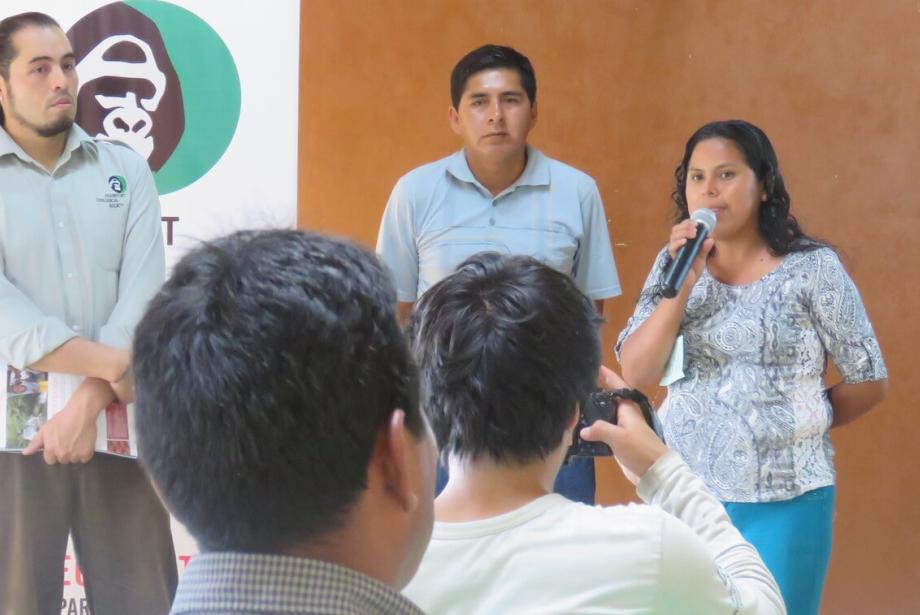 Tres personas de pie frente a un grupo, una mujer hablando por el micrófono.