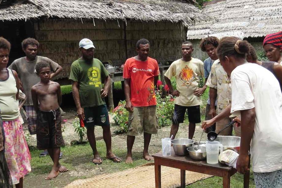Un grupo de hombres y mujeres rodean a una mujer en una pequeña mesa que demuestran cómo presionar el aceite de coco.