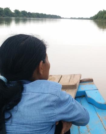 Back of the head of a woman in a boat, looking out over calm, brown river.