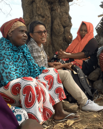 Small group of women, most in bright, patterned clothes, sitting outside, water in background.