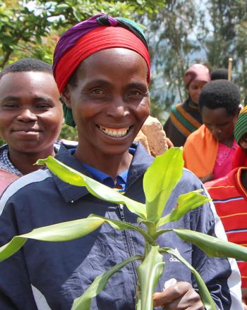 Miembros de la comunidad con ropa brillante. La mujer al frente está sonriendo y sosteniendo la planta.