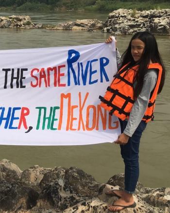 Two girls standing in front of river holding hand-made sign that says "Lancang-MEkong is the Same River We Share the Same Mother, the Mekong"