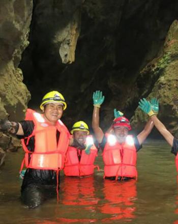 Cinco personas con cascos y chalecos naranjas en una cueva, con el agua a la altura de la cintura, sonriendo y saludando a la cámara.
