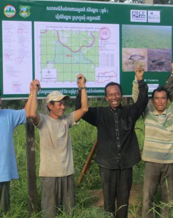 Grupo de 5 hombres parados afuera, tomados de la mano en el aire y sonriendo frente a un cartel con un mapa.