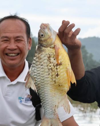 Two smiling men sitting in a boat, with pne holding a Julliens Golden Carp fish.