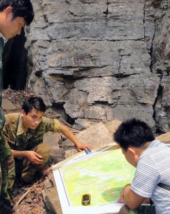 Four men, outside, bent over looking at large map laid out on a rock.